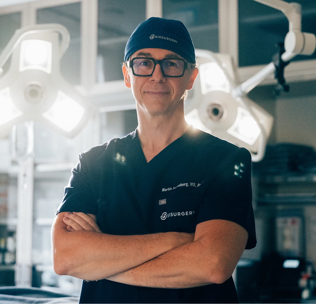 Portrait of Dr. Martin Jugenburg in dark blue scrubs and a surgical cap, standing with his arms crossed in a modern operating room with bright surgical lights in the background.