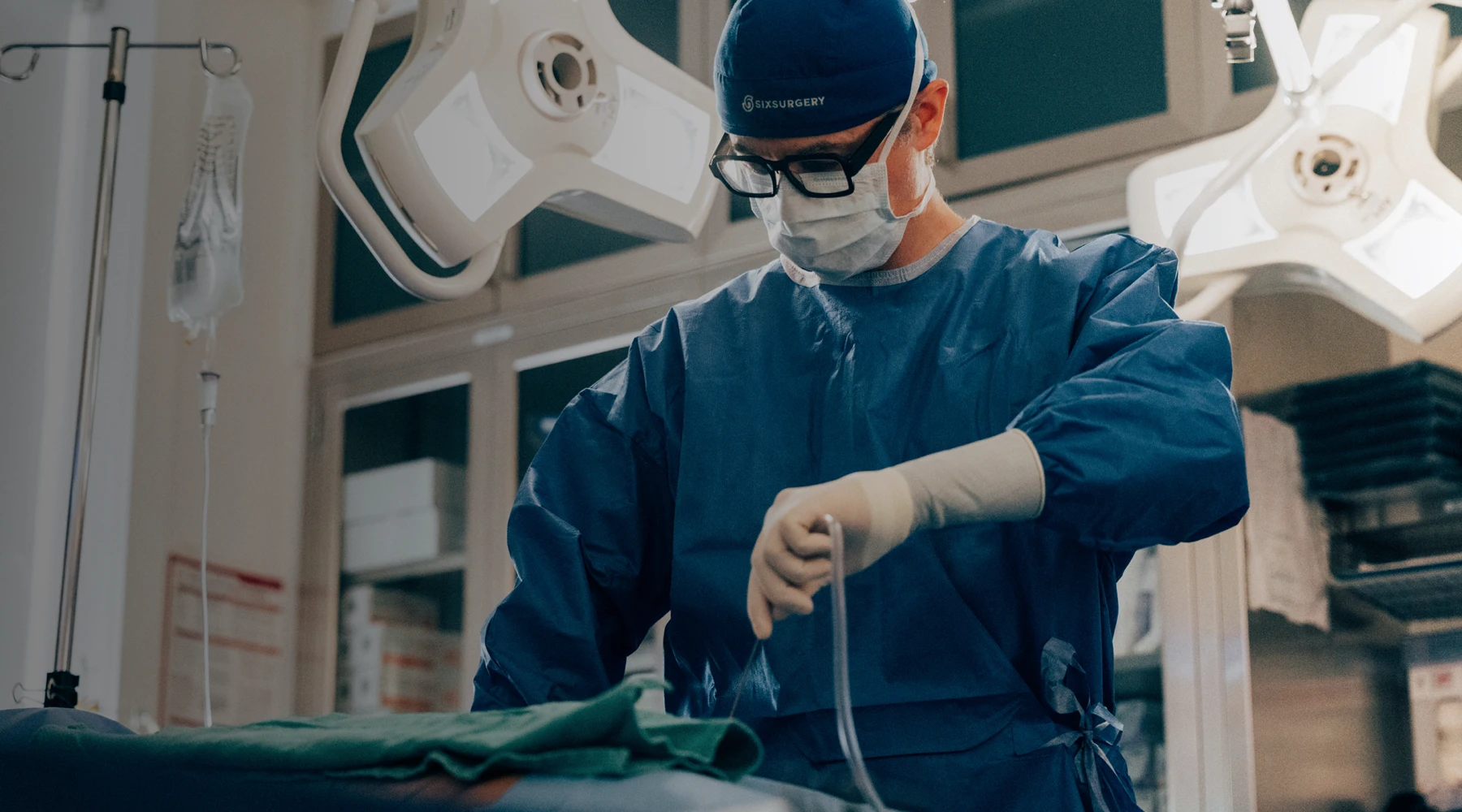 Dr. Martin Jugenburg in full surgical attire, including a face mask and sterile gloves, performing a procedure in a brightly lit operating theater.