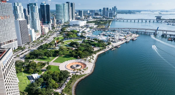An aerial panoramic view of a coastal city skyline featuring high-rise buildings, a lush green waterfront park, and a harbor with several boats.