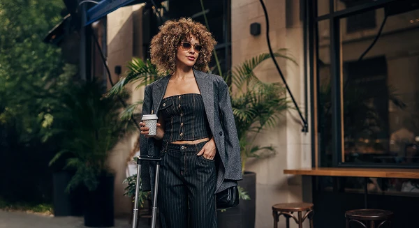 A stylish woman with curly hair wearing sunglasses and a pinstripe suit standing with a rolling suitcase and a coffee cup in a lush, outdoor urban setting.
