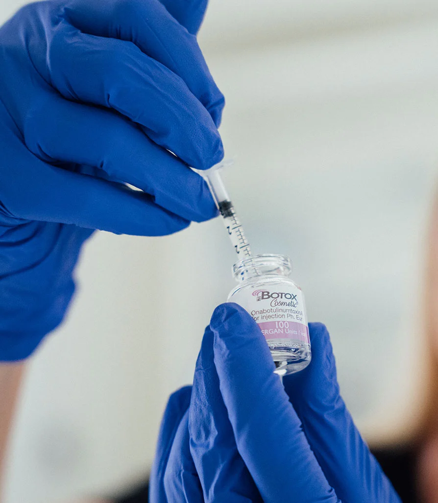 Close-up of hands in blue medical gloves using a syringe to draw liquid from a glass vial labeled "Botox Cosmetic."