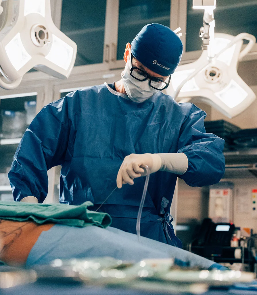 A surgeon wearing a blue cap, mask, and glasses focused on performing a surgical procedure in a sterile, brightly lit operating room.