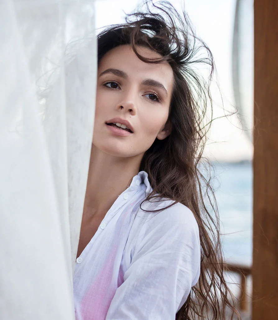 A woman with long dark hair peeking through white curtains, looking off-camera with a soft, elegant expression.