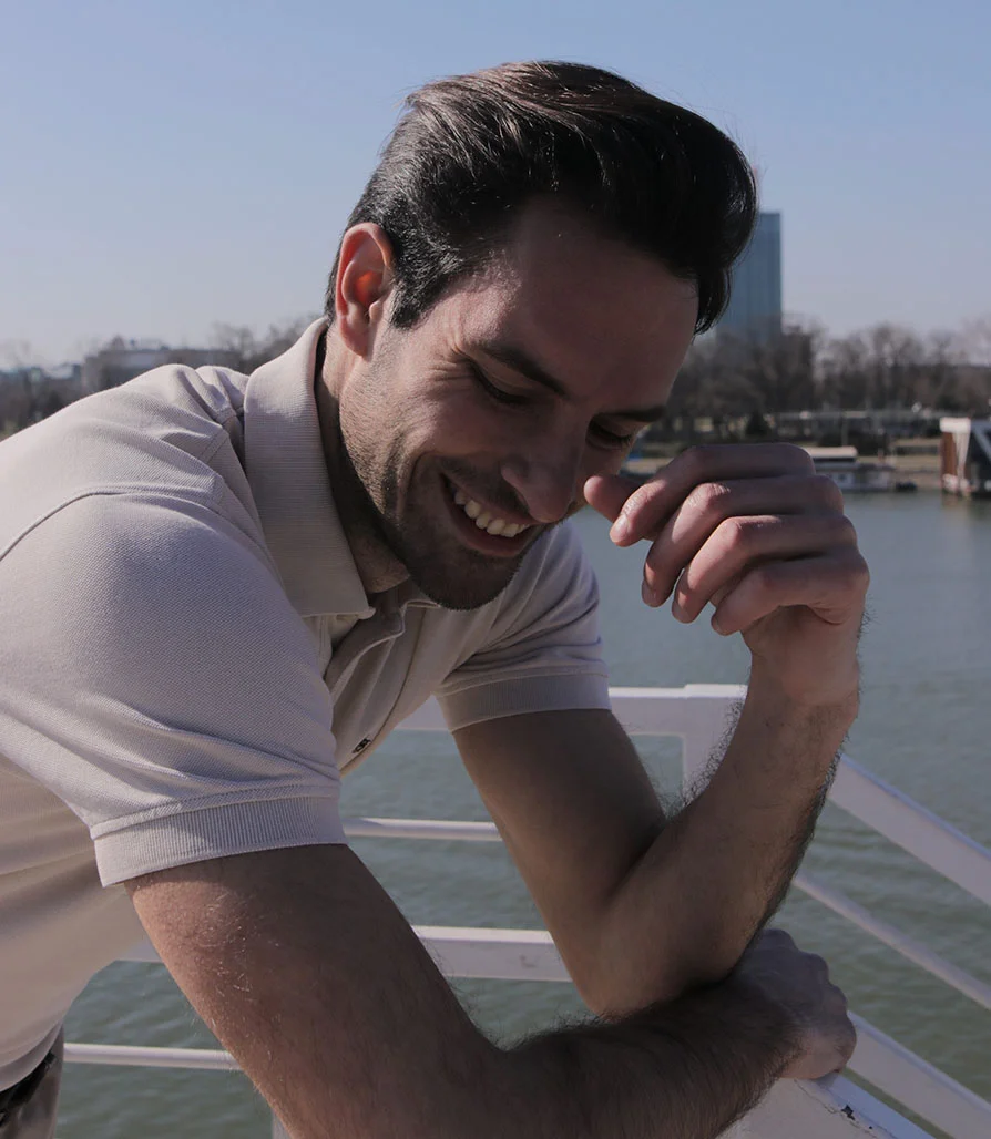A man with dark hair in a beige polo shirt smiling and leaning on a white railing with a river in the background.
