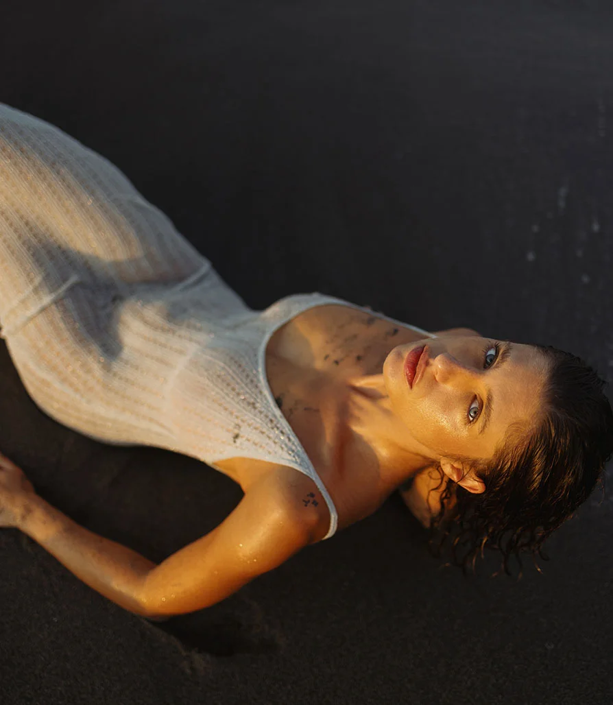 A high-fashion overhead shot of a woman on dark sand, displaying glowing, healthy skin resulting from a professional skincare regimen.