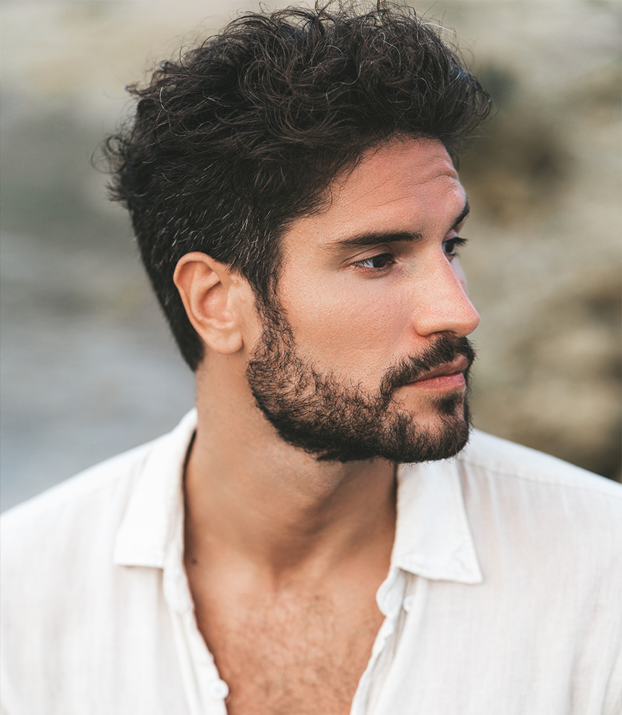 Close-up profile of a man with a thick, well-groomed beard and curly dark hair, wearing a white linen shirt against a soft outdoor background.