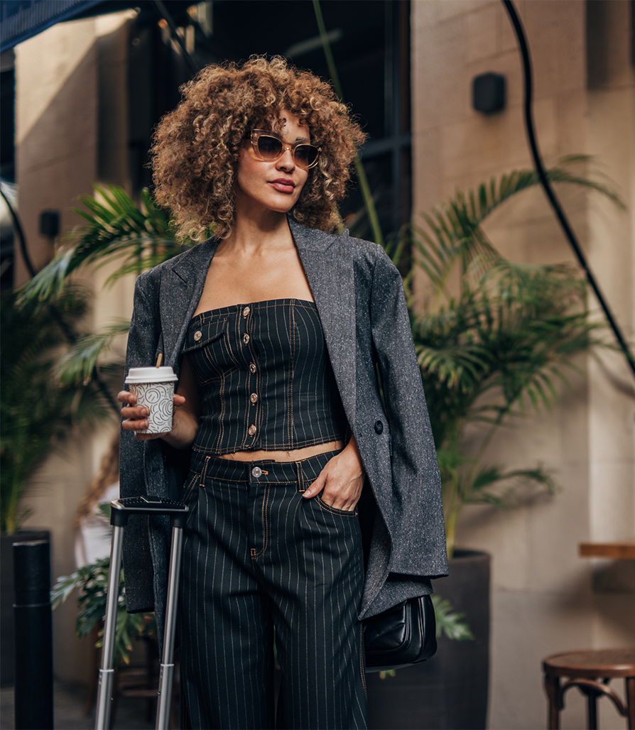 A stylish woman with curly hair wearing sunglasses and a pinstripe suit standing with a rolling suitcase and a coffee cup in a lush, outdoor urban setting.